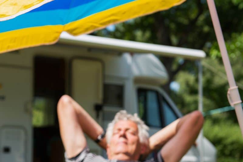 Mann beim Camping unter Sonnenschirm, im Hintergrund Wohnmobil